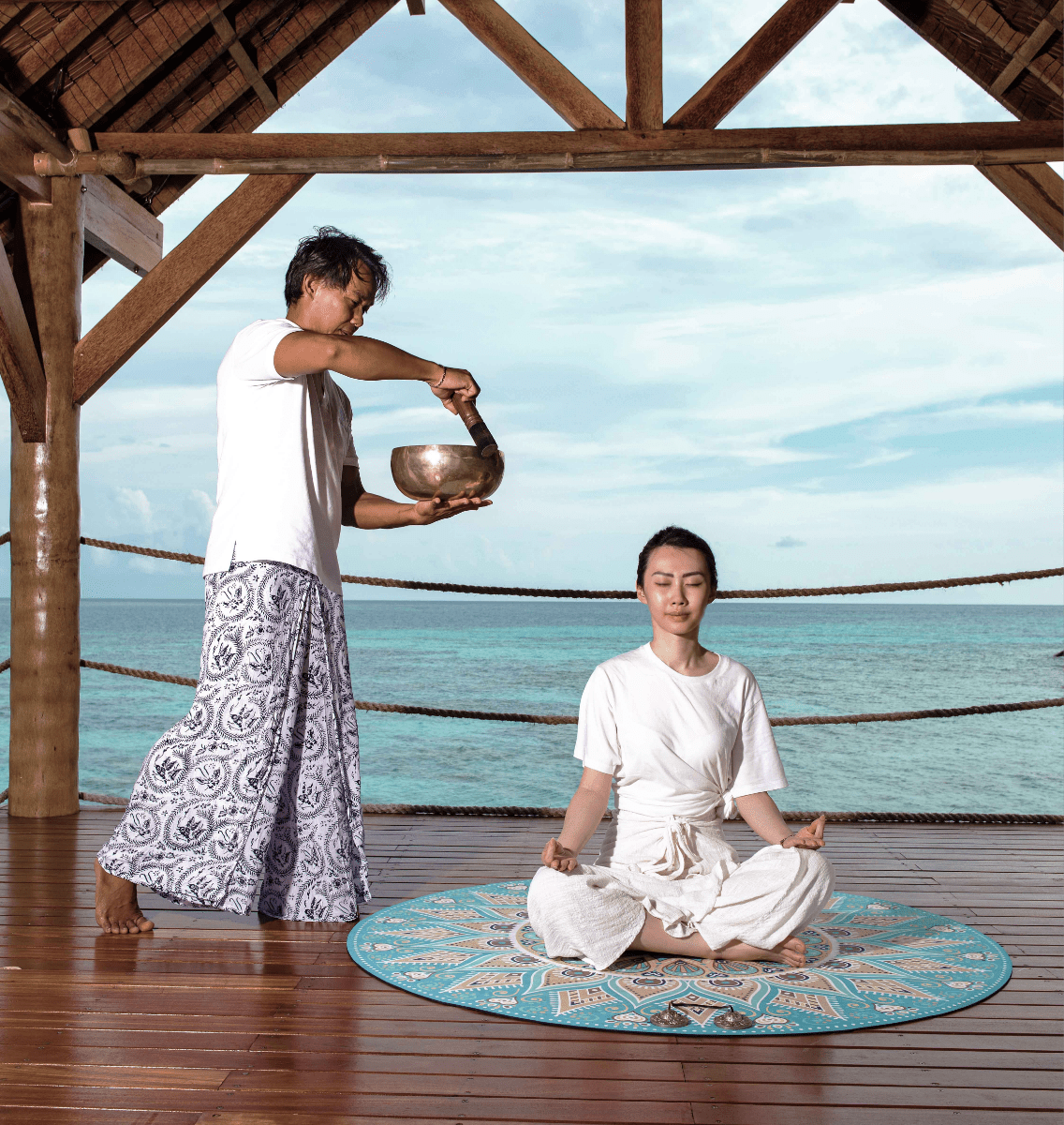 HR-woman-meditating-elang-wooden-deck-male-staff-holding-bowl-lagoon-behind-2